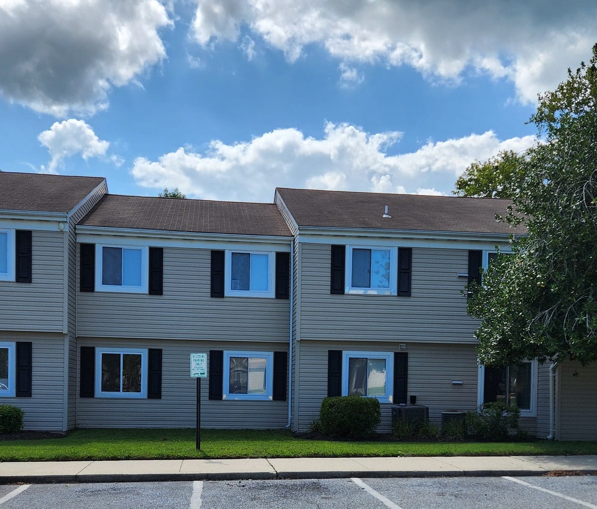 A two-story tan-sided apartment building at Prince Frederick Village under a blue summer sky, with a green lawn and resident parking out front