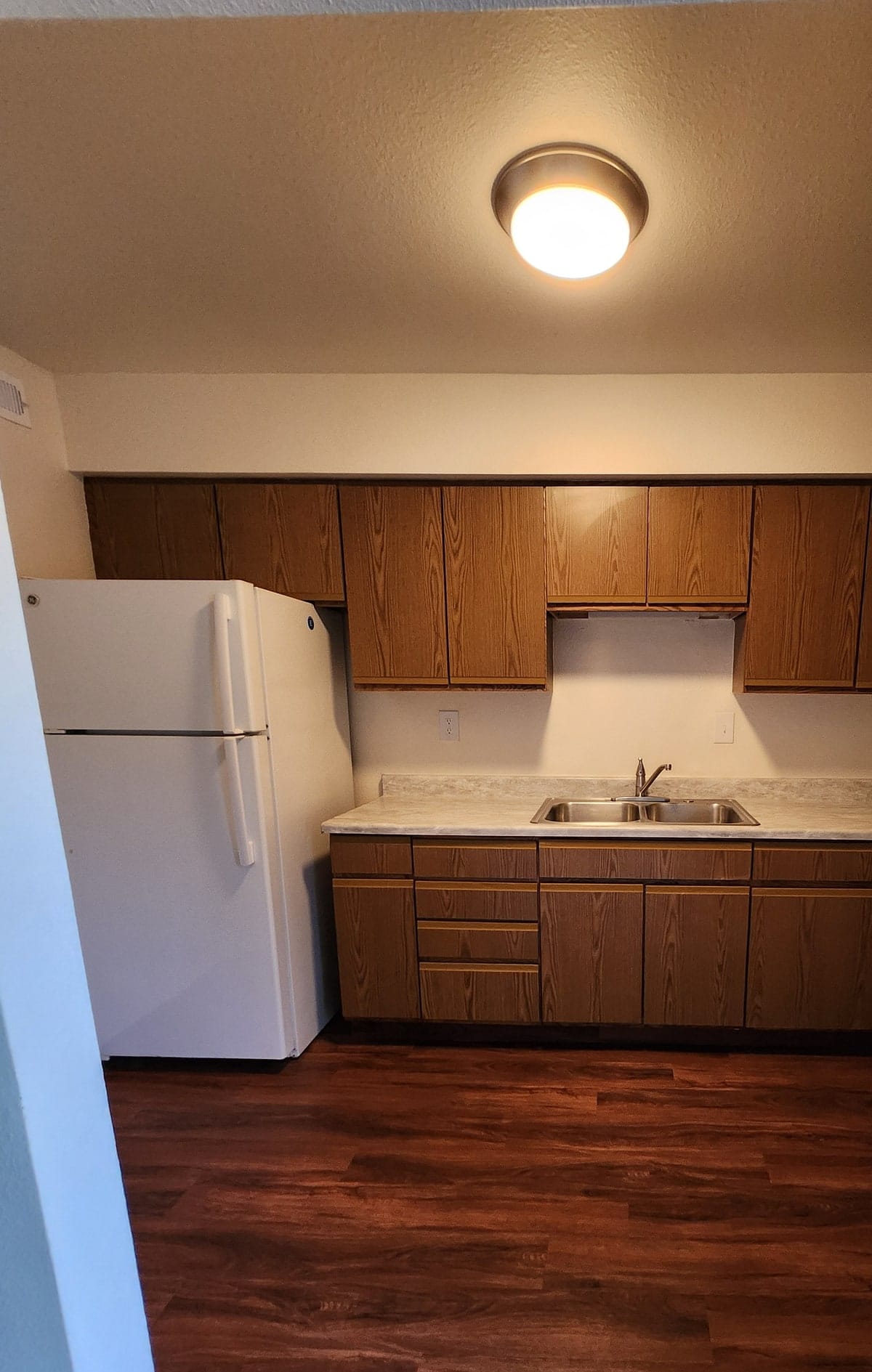 A galley kitchen at Prince Frederick Village with woodgrain cabinets and a double stainless sink over dark plank flooring, seen from the doorway