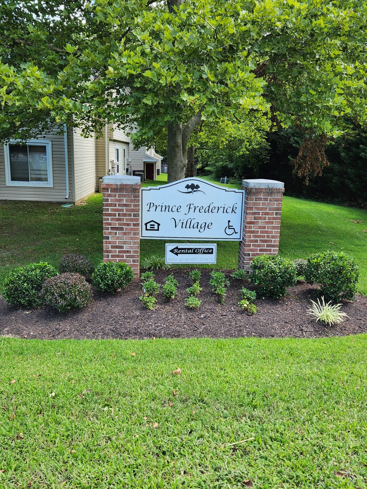The Prince Frederick Village entrance sign on brick piers, set in a mulched garden bed beneath a large shade tree, with a rental-office arrow below